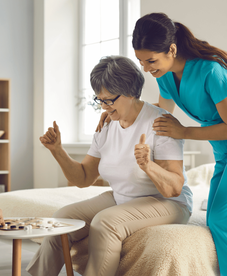 Female caregiver leaning over older female, playing board game, both smiling
